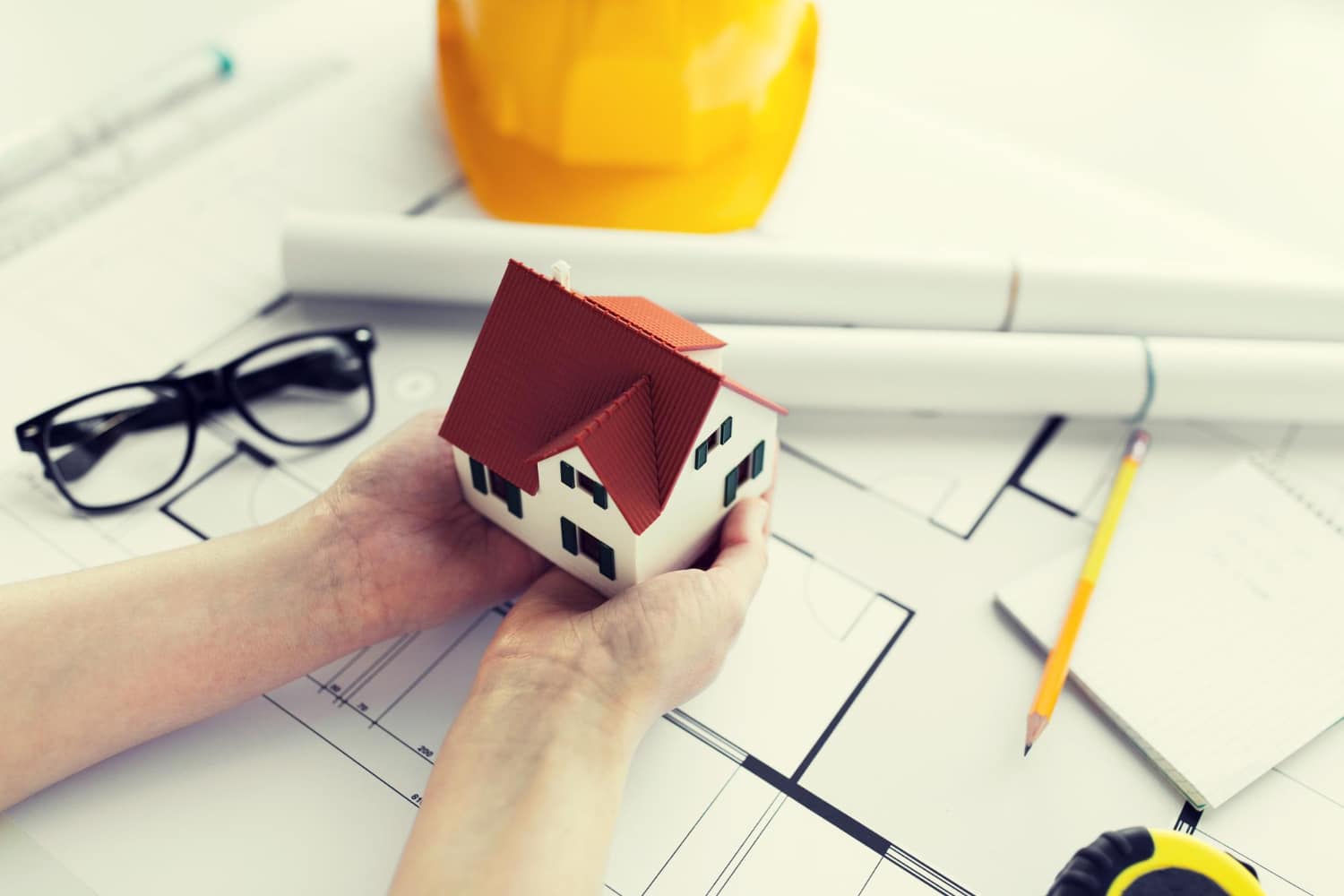un professionnel tient une petite maison dans ses mains, au dessus d'un plan de construction.
